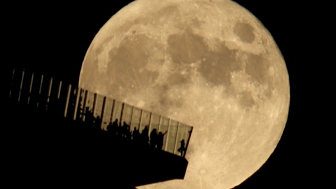 People standing on the Edge NYC outdoor observation deck are silhouetted by the full Hunter's Moon rising in New York City on October 20, 2021 as seen from Hoboken, New Jersey. Earlier in the day Related Companies, the owner of Hudson Yards announced KKR & Co. bought a majority stake in EdgeNYC. (Photo by Gary Hershorn/Getty Images)