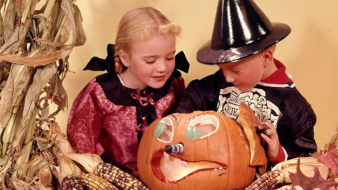 Two children wearing Halloween costumes look inside a carved jack-o'-lantern pumpkin, ca 1960s.