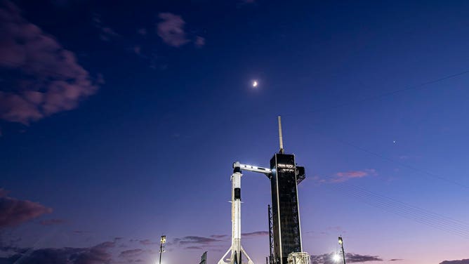 A SpaceX Falcon 9 rocket on Kennedy Space Center launchpad 39A with Jupiter, Saturn, the Moon, above the rocket and Dragon.