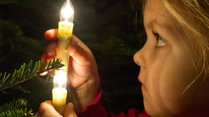 A little girl holds electric lights in the shape of candles while decorating a Christmas tree.