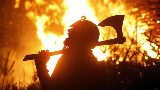 A Viking re-enactor stands in the foreground as a fire burns behind him.