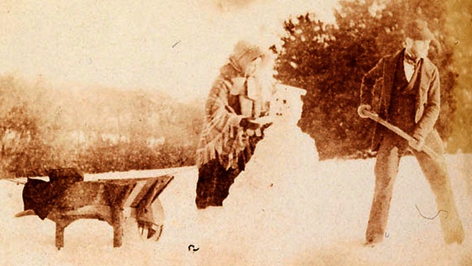 A man shovels snow while a woman places the finishing touches on a snowman. This is one of the earliest photos of a snowman.