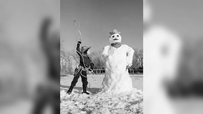 Dressed as a cowboy, a 1950s boy attempts to lasso a snowman that is also dressed as a cowboy.
