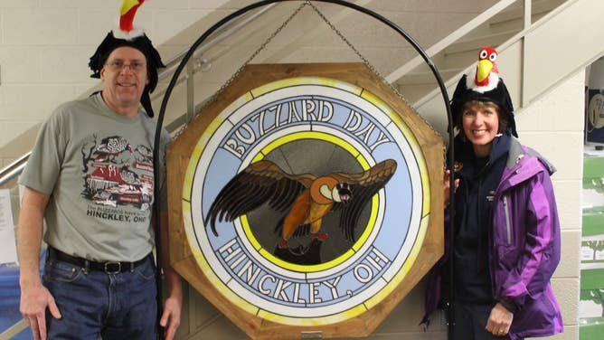 Buzzard Day goers wear buzzard hats as they stand next to a Hinckley, Ohio sign that displays a flying buzzard.