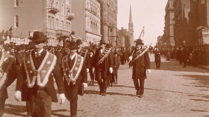 Men march in the Saint Patrick's Day Parade, wearing Irish sashes and carrying banners, in midtown Manhattan, New York City in 1895.