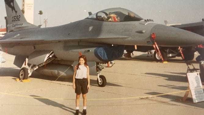 A young Waddington smiles as she stands in front of an F16 aircraft.