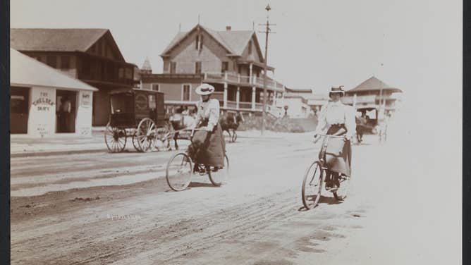 Two women ride bicycles on a dirt road in Long Branch, New Jersey in the late 19th century.