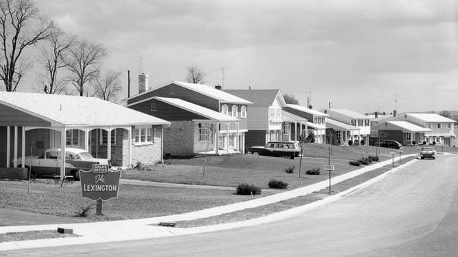 Homes, each with their own lawn, line a neighborhood street in the mid-20th century.