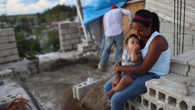 A mother holds her baby as her husband mixes cement and tried to rebuild their home in December 2017