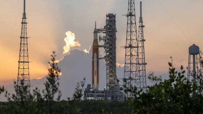 The Space Launch System rocket sits on the launch pad. In the background, the sun rises behind clouds. Green bushes sit in the foreground.