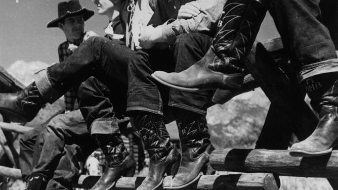A line of cowboys resting on a fence and showing off their boots. Circa 1955.