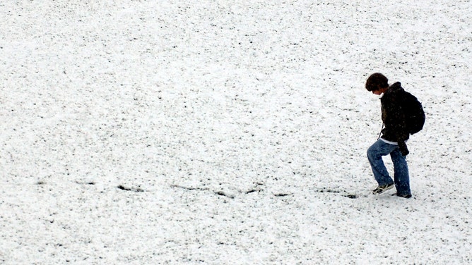 A young man walks backwards across the field below the school so he can see his footprints in the snow. 