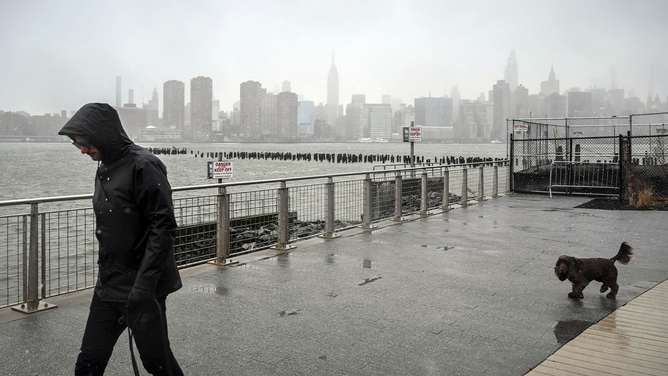 A person walks during rain and snow before the New York City skyline in Brooklyn on March 14, 2023. - A major Nor'easter is expected to bring heavy snow and strong winds to the East Coast on Wednesday, forecasters say.