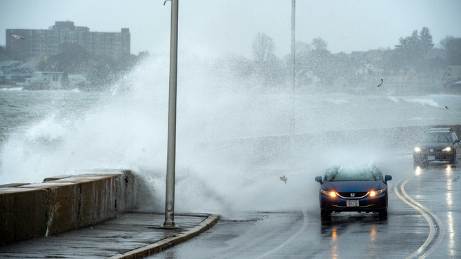 Waves crash over the sea wall and onto cars as they drive in Winthrop, Massachusetts, on March 14, 2023. - The noreaster storm brought high winds that created large waves which caused minor costal flooding.