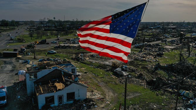 Crews work to clear the land under a giant American flag that flies on a crane in the midst of destruction wrought by a massive tornado that laid waste to much of the town of Rolling Fork, Mississippi, on March 28, 2023.