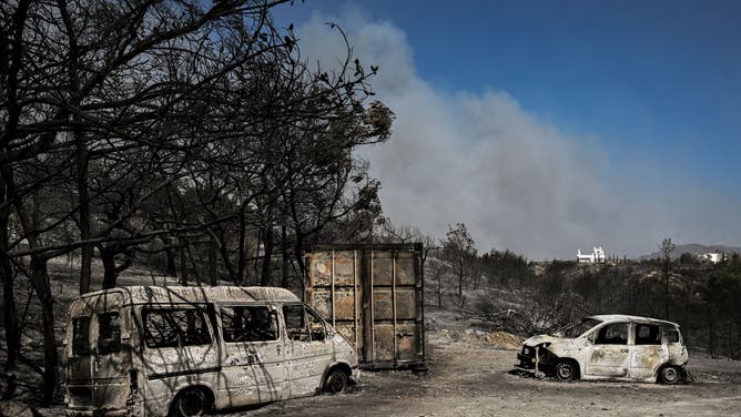 This photograph taken on July 24, 2023, shows burnt vehicles and smoke in the background during a fire between the villages of Kiotari and Genadi, on the Greek island of Rhodes. Firefighters tackled blazes that erupted in peak tourism season, sparking the country's largest-ever wildfire evacuation -- and leaving flights and holidays cancelled. Tens of thousands of people have already fled blazes on the island of Rhodes, with many frightened tourists scrambling to get home on evacuation flights.