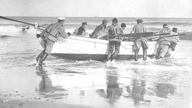 Nauset Life-Saving crew launching their surfboat, all wearing mixed uniforms with wading boots and cork life vests; no date, possibly post-1915 era.
