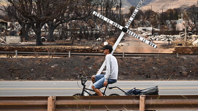 A man on a bike carries a cross in the north side of Lahaina, Hawaii, on August 16, 2023.