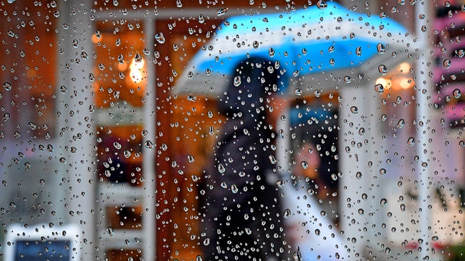 FILE - People take shelter from the rain under umbrellas as they are viewed through the raindrops collected on an automobile window in Sayville, New York, on Feb. 10, 2020.