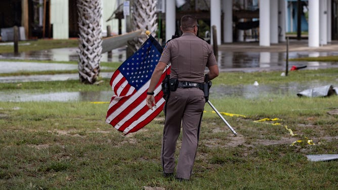 A sheriff's deputy picks up an American flag from debris following Hurricane Idalia in Horseshoe Beach, Florida, US, on Wednesday, Aug. 30, 2023. Hurricane Idalia knocked out power to hundreds of thousands of Florida customers, grounding more than 1,800 flights and unleashing floods along far from where it came ashore as a Category 3 storm earlier Wednesday. Photographer: Christian Monterrosa/Bloomberg