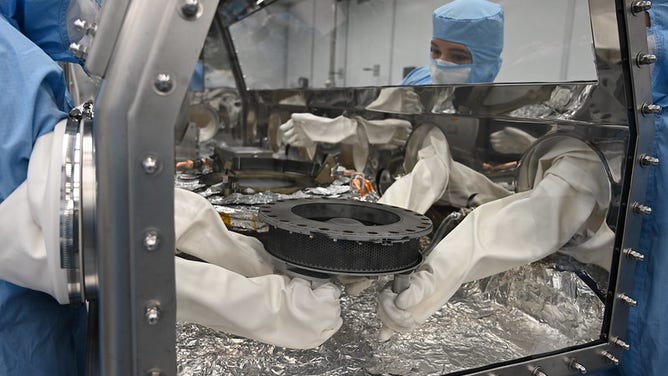 Astromaterials processor Mari Montoya and OSIRIS-REx curation team members set the TAGSAM (Touch and Go Sample Acquisition Mechanism) down in the canister glovebox after removing it from the canister base and flipping it over.