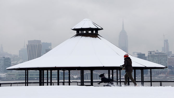 A man walks his dog in a snow-covered park in front of the skyline of midtown Manhattan and the Empire State Building in New York City on February 17, 2024, in Hoboken, New Jersey.