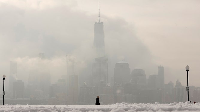 A woman walks through a snow-covered park in front of the skyline of lower Manhattan and One World Trade Center in New York City on February 17, 2024, in Hoboken, New Jersey.