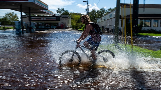 A man rides his bicycle through water as roads and businesses are flooded by the rising Anclote River during the recovery from Hurricane Milton on October 11, 2024 in New Port Richey, Florida.