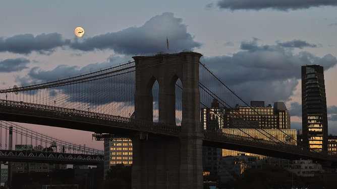 A 96% illuminated Hunter's Moon rises above the Brooklyn Bridge as the sun sets on October 15, 2024, in New York City.