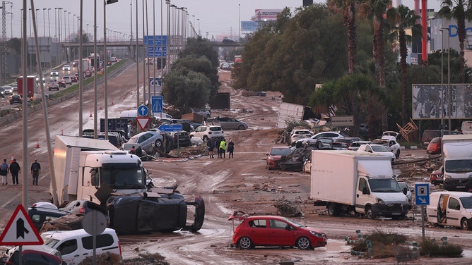 2024年11月1日、スペイン・バレンシア郊外のマサナッサ市近くのV-31高速道路沿いで、最近の洪水の残骸に巻き込まれた車やトラックの前を歩く人々。