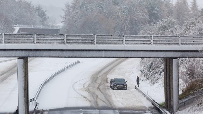 スターリング近郊の雪に覆われた空いている M9 高速道路で、車の隣に男性が立っています。ストーム・バートが強風、大雨、雪、氷で国を襲っている。黄色の警告が発効し、その結果、 "生命と財産に対する潜在的なリスク"。撮影日：2024年11月23日（土）