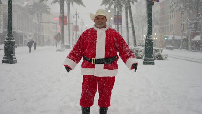 A man dressed in a cowboy hat and Santa outfit, with snow-covered palm trees behind him in New Orleans.