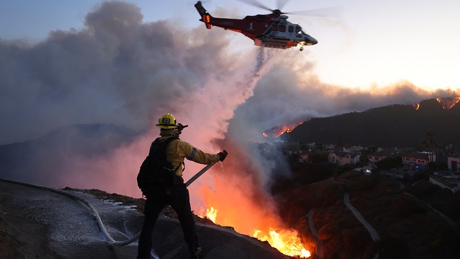 Fire personnel respond to homes destroyed while a helicopter drops water as the Palisades Fire grows in Pacific Palisades, California on January 7, 2025. A fast-moving wildfire in a Los Angeles suburb burned buildings and sparked panic, with thousands ordered to evacuate January 7, 2025 as "life threatening" winds whipped the region.