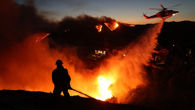 Fire personnel respond to homes destroyed while a helicopter drops water as the Palisades Fire grows in Pacific Palisades, California on January 7, 2025.