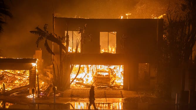 カリフォルニア州ロサンゼルス – 1月8日: カリフォルニア州ロサンゼルスで2025年1月8日、強力なハリケーンの最中、パシフィック・コースト・ハイウェイ沿いの家々を焼くパリセーズ・ファイアの炎を見守る消防士。南カリフォルニアのサンタアナの強風と乾燥した状況の中、急速に進行する山火事が2,900エーカー以上に広がり、沿岸地域の住宅を脅かしている。