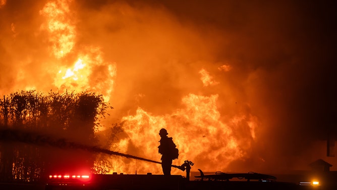 2025年1月8日、カリフォルニア州ロサンゼルスで、強力なハリケーンの最中、パシフィック・コースト・ハイウェイ沿いの家々を焼くパリセーズ火災を消火するために消防車の上に立つ消防士。