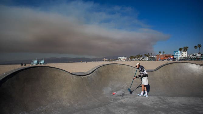 Skateboarder Haden Mckenna sweeps ashes from the Venice Skatepark from the Palisades Fire wildfire to skate as smoke rises from the Palisades Fire wildfires in the background on January 11, 2025 in Los Angeles, California. 