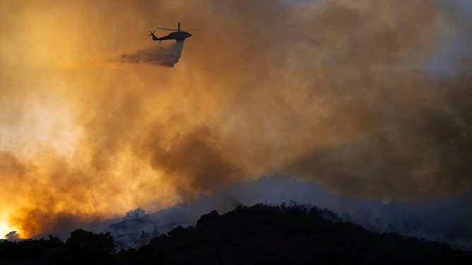 A helicopter makes a water drop on fire that encroaches on a home in the Mandeville Canyon area where homes were evacuated on Saturday, January 11, 2025.