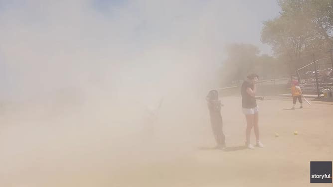 Softball players and coaches cover their eyes as a dust devil overtakes a softball field.
