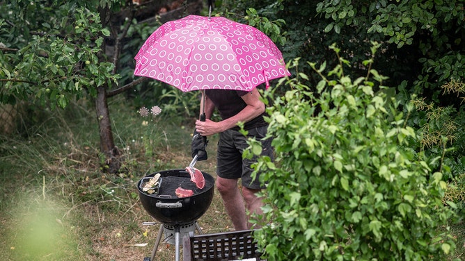 A man, caught in a rainstorm, stands in his backyard at a charcoal grill, turning meat on the grate.