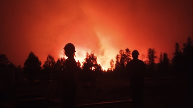 In this handout image provided by Grand Canyon National Park, Two firefighters stand silhouetted against an intense orange-red sky as the Dragon Bravo Fire burns through a forested area at night, with flames and smoke rising in the background at Grand Canyon National Park on July 12, 2025.