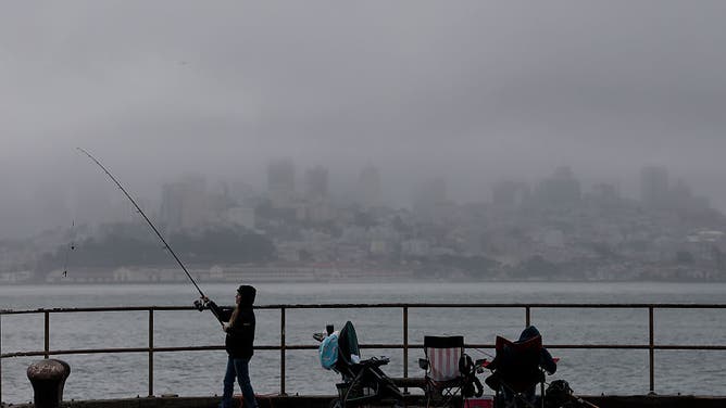 SAUSALITO, CALIFORNIA - JULY 21: Fog obscures the San Francisco skyline as a fisherman prepares to cast a line on July 21, 2025 in Sausalito, California. As summer temperatures soar to record highs across much of the U.S., San Francisco is experiencing a very mild summer as a persistent marine layer hangs over the city on most days, keeping daily temperatures in the 50s and 60s. Since May, the city has only once reached a daytime high temperature of 70 degrees. 