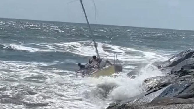 People on the sailboat cling to its side as it hits the rocks and starts tipping.
