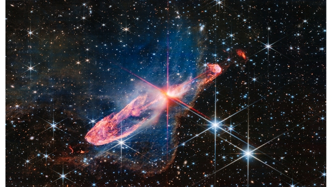 At the center is a thin horizontal orange cloud known as Herbig-Haro 46/47 that is uneven with rounded ends, and tilted from bottom left to top right. It takes up about two-thirds of the length of this angle, but is thin at the opposite angle. At its center is a set of very large red-and-pink diffraction spikes in Webb’s eight-pointed pattern. The vertical spikes extend almost to the top and bottom of the frame. The smaller, fainter diffraction spikes in the center are aligned at a true horizontal, but one set of the longer, more opaque spikes run along the orange cloud. At the middle of the diffraction spikes is a yellow-white blob, which hides two tightly orbiting stars.