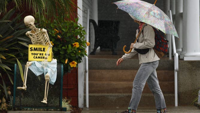 Encinitas, CA - October 14: A woman walks in the rain along Coast Highway as a skeleton is displayed outside of a shop on October 14, 2025 on Encinitas, CA.