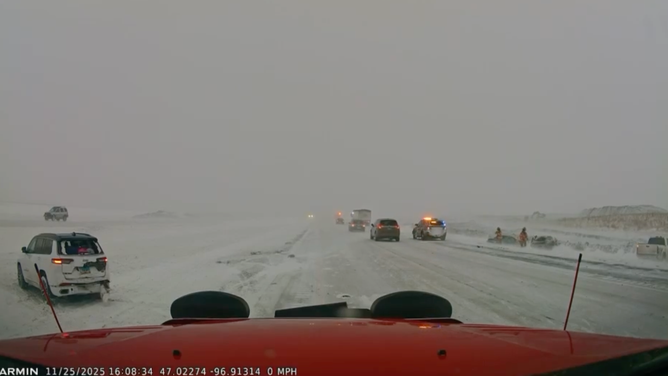 Vehicles slowly drive through the blizzard with hazards past the crash site.