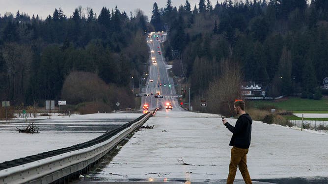 A person stops to capture cellphone images as flood waters from the Snohomish River cover a portion of State Route 9 in Snohomish, Washington, on December 11, 2025. Tens of thousands of people were under evacuation orders Thursday in western North America, after days of heavy rain forced rivers to burst their banks. Storms have battered Washington state in the US and British Columbia over the Canadian border for several days, with rivers continuing to rise.