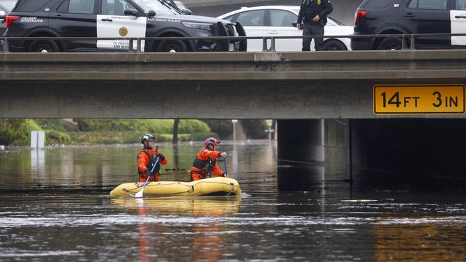 San Diego, CA - January 1: A San Diego Police officer, standing on State Route 163, and members of the San Diego Lifeguard River Rescue Team look for people after the San Diego River overflowed and flooded areas in Mission Valley during a storm on January 1, 2026 in San Diego, CA.