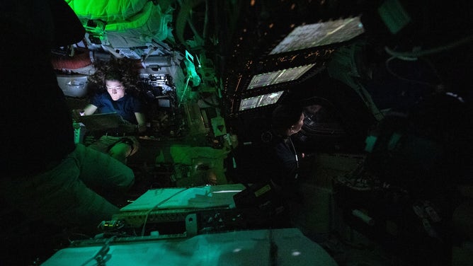 Astronaut Christina Koch is nestled into a corner of the Orion crew capsule, reading. The rest of the capsule is dark, but her face it lit by the glow from her tablet. Two of her other crew members can dimly be seen looking out the window and floating in the foreground