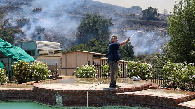 Simi Valley, CA - May 18: Barton Richter hoses down his backyard on Rocking Horse Dr. as the Sandy Fire burns nearby in Simi Valley, CA on Monday, May 18, 2026.
